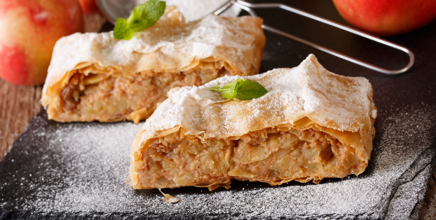 Zwei Stücke Apfelstrudel mit Puderzucker und Minzblättern auf einer dunklen Platte.