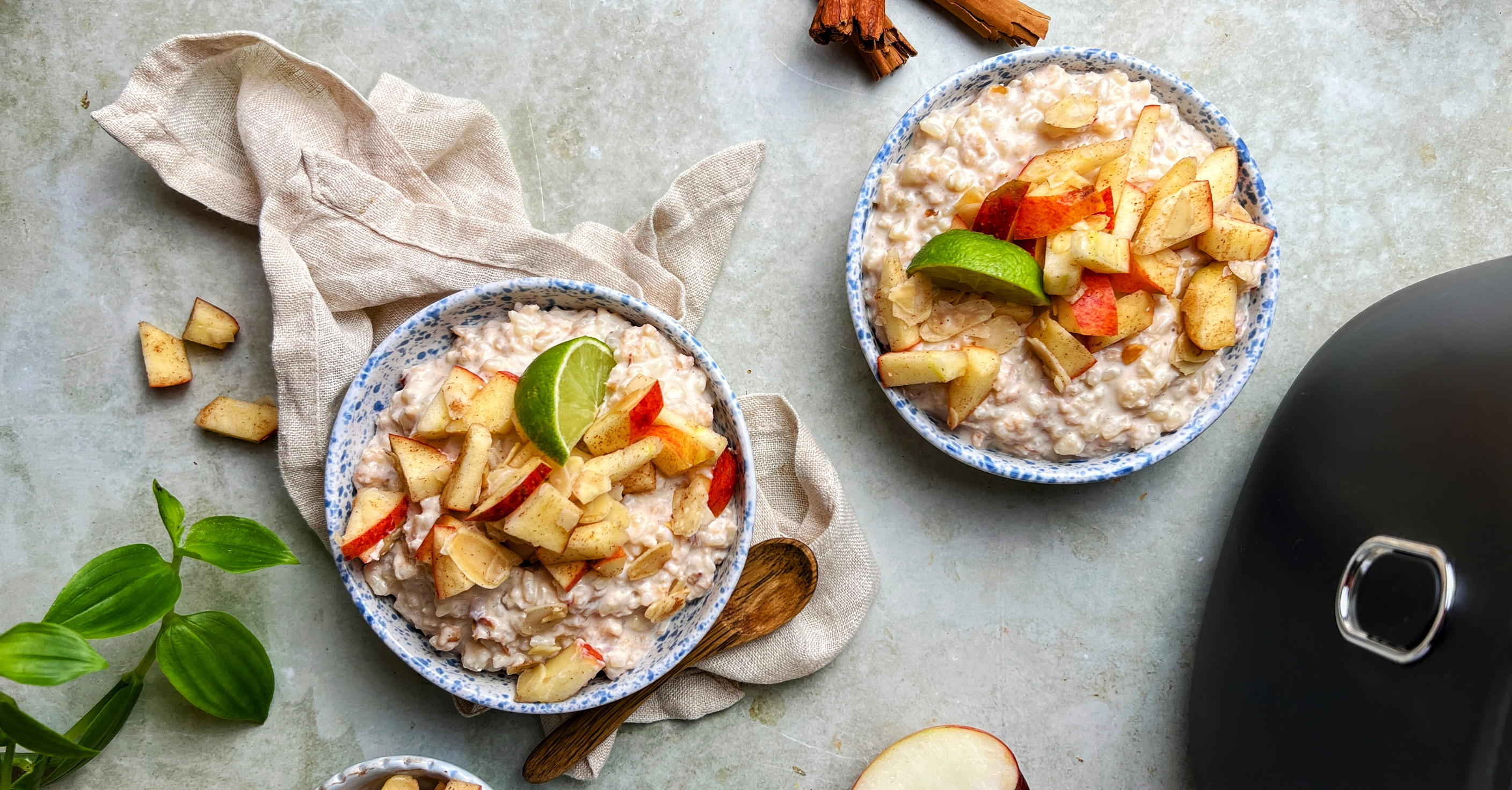 Bols de porridge crémeux avec morceaux de pomme sur une surface claire, accompagnés d’un linge et d’ustensiles.